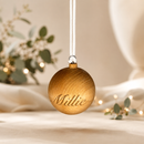 Wooden ornament with 'Millie' engraved, hanging against a soft, blurred background with lights and greenery.