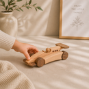 Wooden toy car being held by a hand on a soft surface with a plant and framed picture in the background.