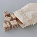 A set of wooden blocks with numbers and basic math symbols, spilling out of a cotton bag onto a gray surface.