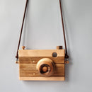 A wooden toy camera with a brown strap, lens, and wooden body, displayed against a grey background.