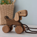 A wooden pull-along dog toy with wheels and a rope attached to it, placed on a floor next to a wall and a basket.