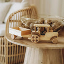 A natural wood toy tractor with a small attached wooden trailer, displayed on a round wooden table in a softly lit indoor setting with a woven chair and glass bowls in the background.