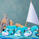 Close-up styled scene with the Bumbu Toys white sailing boat behind teal wooden waves, three swans and rocks.