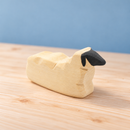 Resting Bumbu wooden sheep with cream carved body and charcoal face on a natural pine surface.